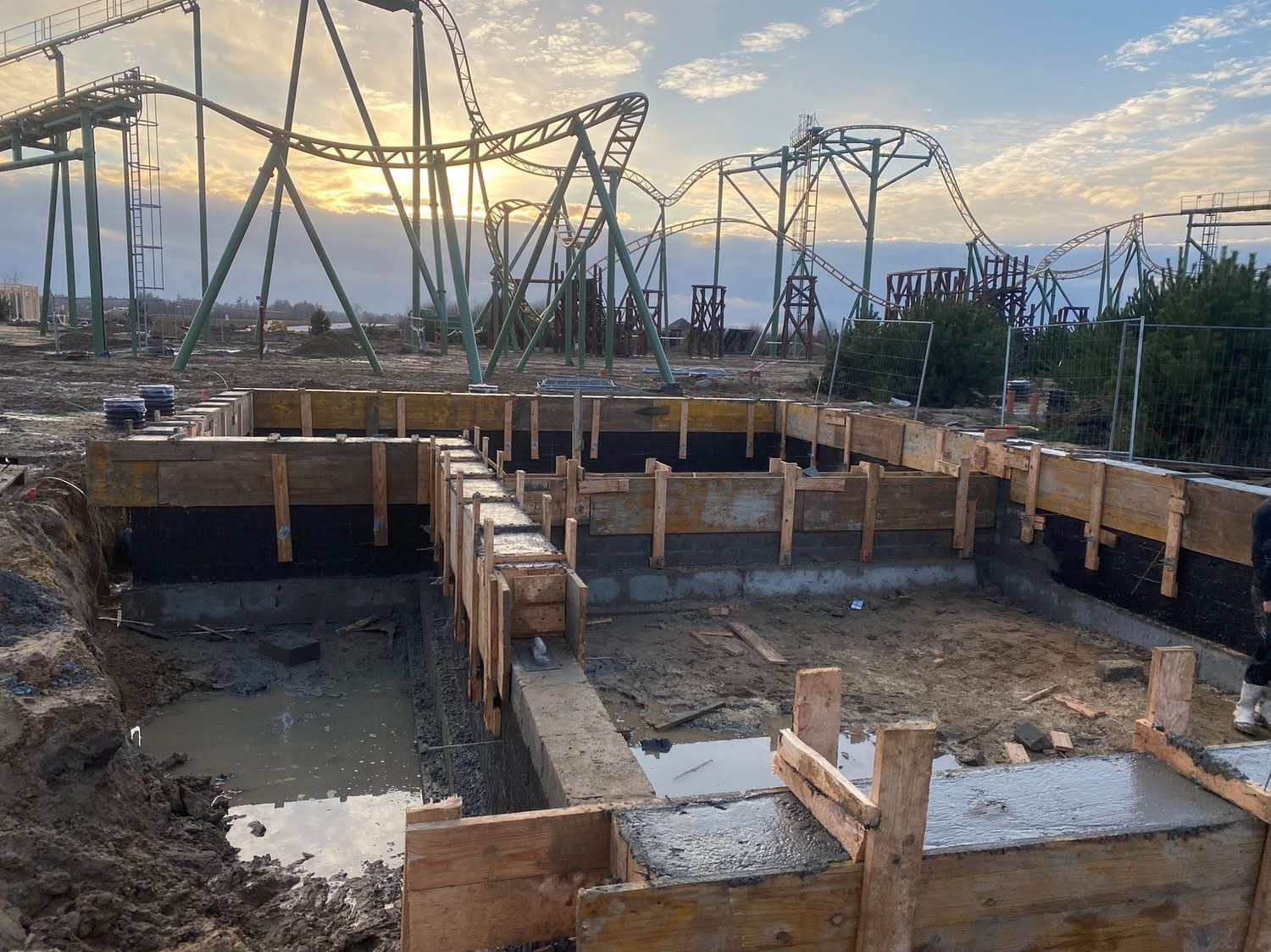 Fundamenty pod budowlę z drewnianym szalunkiem, częściowo zalane betonem. W tle konstrukcja kolejki górskiej i zachodzące słońce.