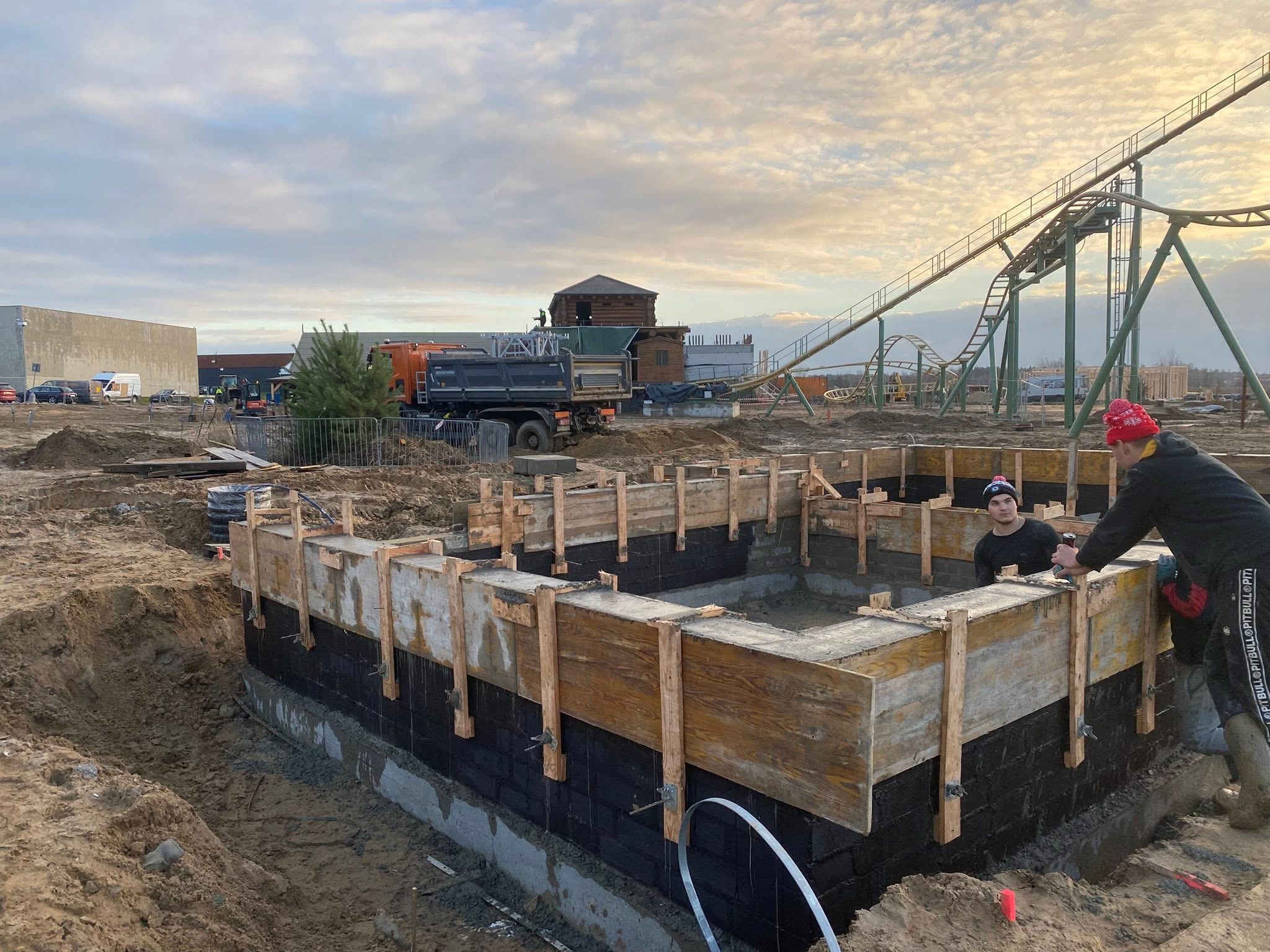 Budowa fundamentów z drewnianym szalunkiem i izolacją bitumiczną, w tle plac budowy z ciężarówką i konstrukcją kolejki górskiej. Dwóch pracowników przy pracy.