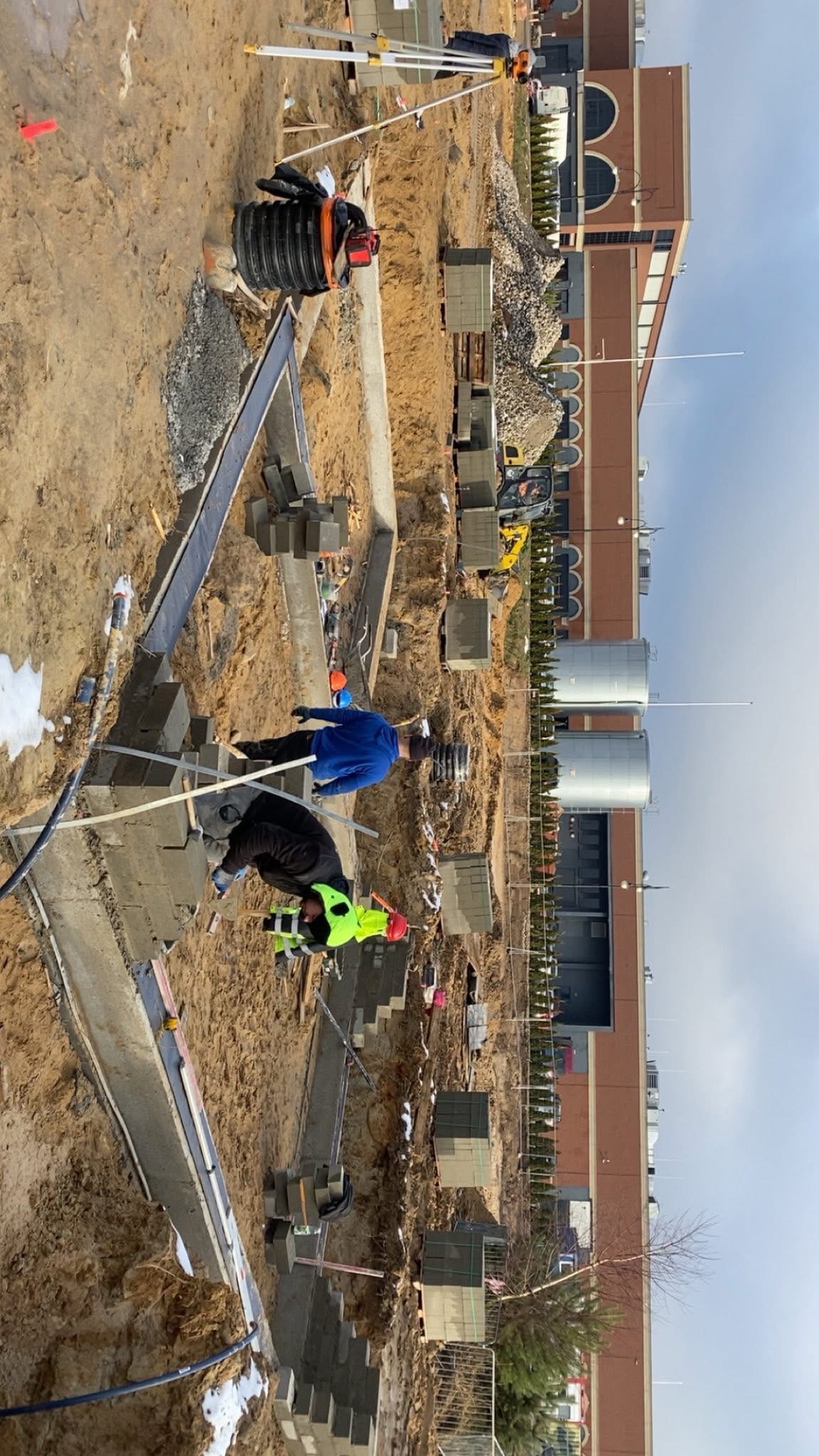 Dwóch murarzy w trakcie wznoszenia fundamentów z bloczków betonowych. Widoczne narzędzia, wykop i częściowo ukończona konstrukcja. W tle budynek i sprzęt budowlany.