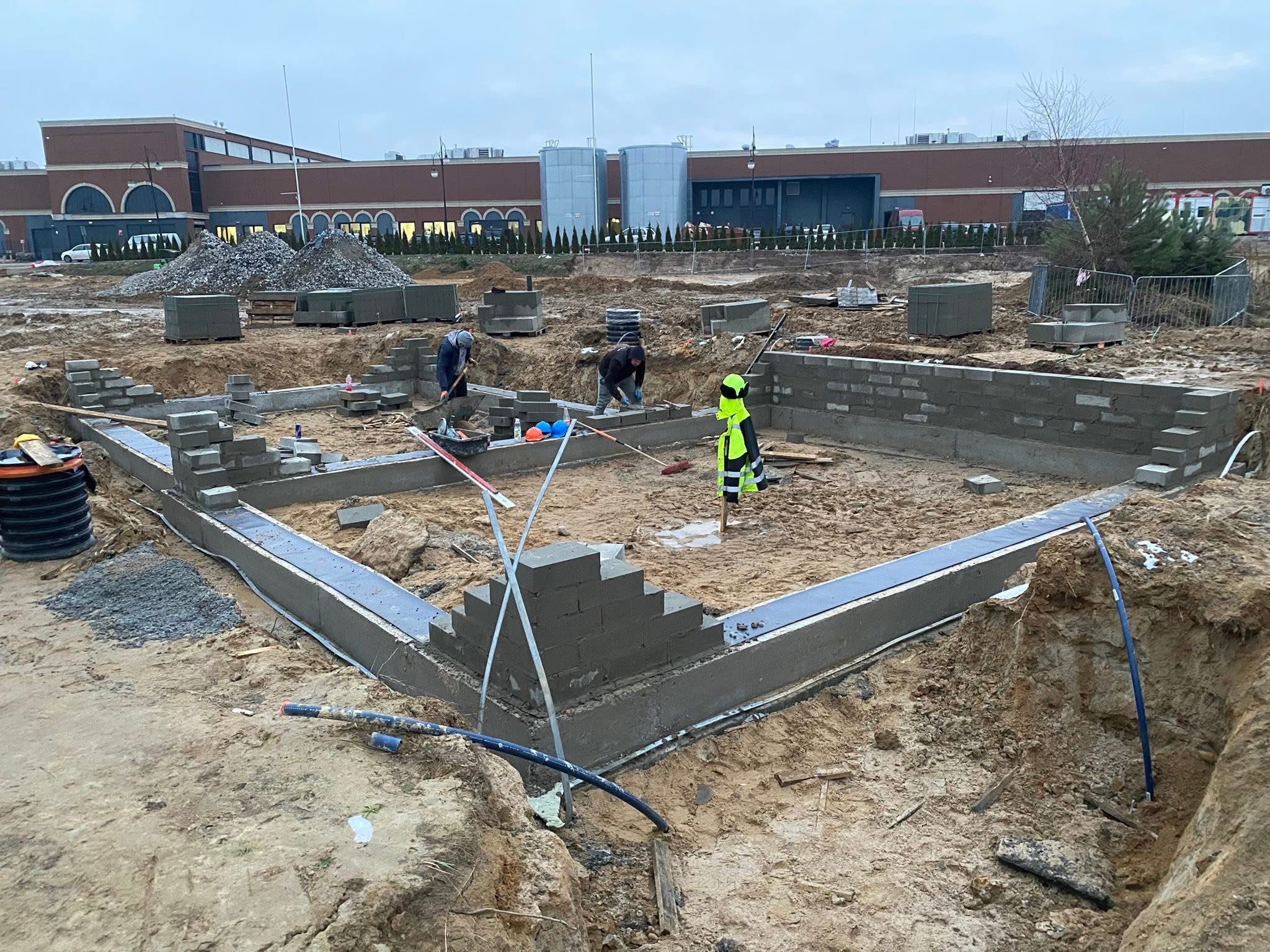 Wykop pod fundamenty z częściowo wymurowanymi ścianami z szarych bloczków. W tle pracownicy budowlani i budynek handlowy w pochmurny dzień.