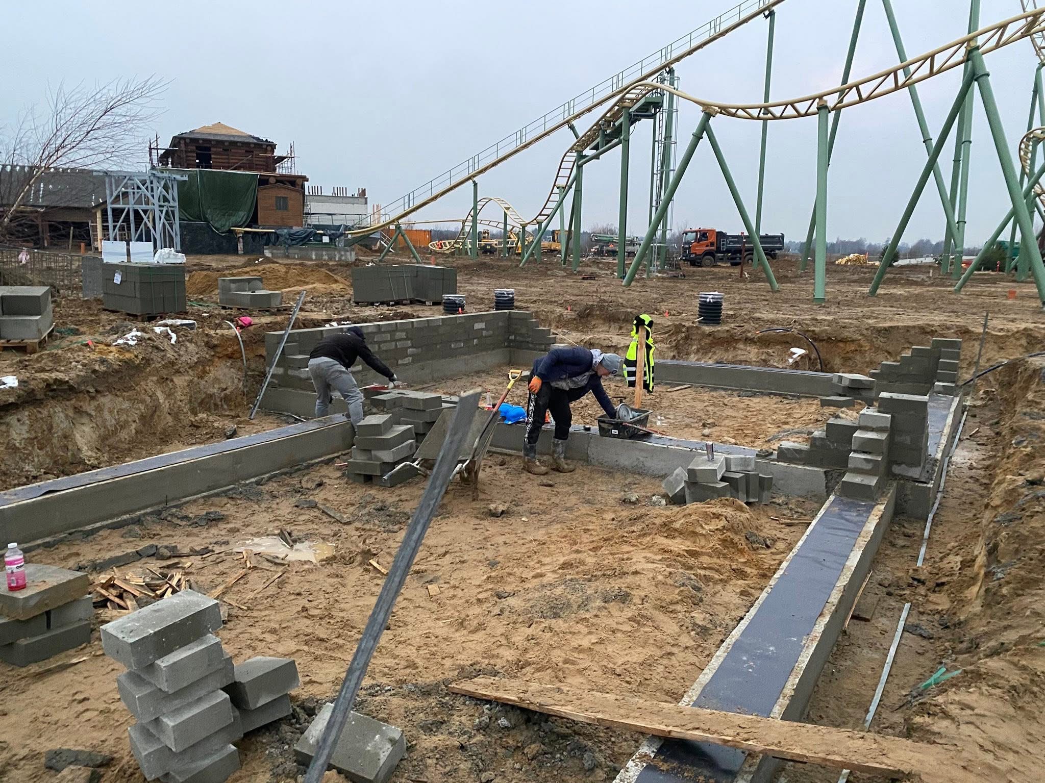 Budowa fundamentów z bloczków betonowych na placu budowy z atrakcjami parku rozrywki w tle. Dwóch pracowników przy pracy, widoczne narzędzia i materiały budowlane.