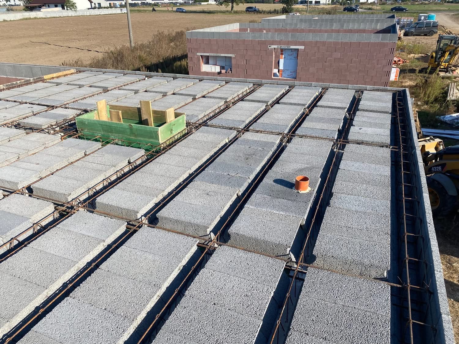 Wieniec stropowy z szalunkiem i zbrojeniem na budowie domu jednorodzinnego. Widoczne bloczki szalunkowe i elementy instalacji. W tle budynek w stanie surowym otwartym.