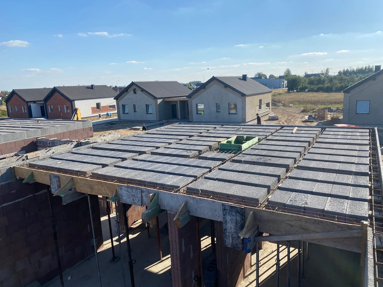 Budowa stropu Teriva z widocznymi belkami, pustakami i zbrojeniem. W tle widać nowo wybudowane domy jednorodzinne pod błękitnym niebem.
