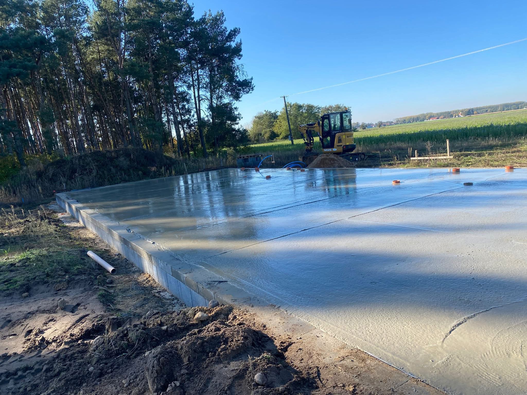 Świeżo wylana, gładka płyta fundamentowa z widocznym obrysem ścian, na tle zieleni i błękitnego nieba, z małą koparką w tle. Widoczne pomarańczowe znaczniki.