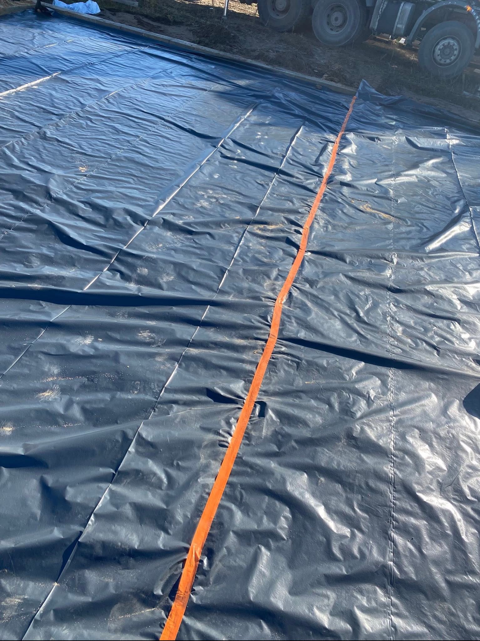 Czarna folia budowlana rozłożona na ziemi, zabezpieczona pomarańczową taśmą. W tle widoczne koła ciężarówki, sugerujące przygotowanie terenu pod budowę.