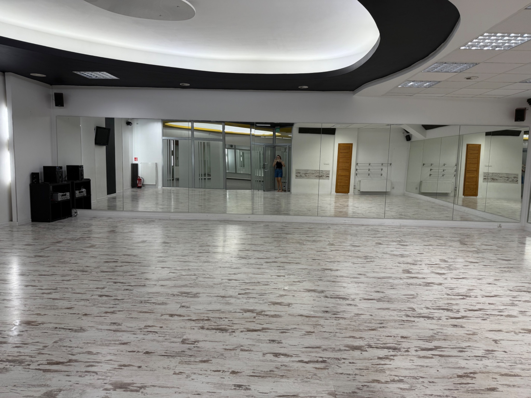 Jasne studio fitness z lustrami, parkietem i czarnym sufitem. W lustrze odbija się osoba robiąca zdjęcie. Widoczny sprzęt audio. Minimalistyczny design.