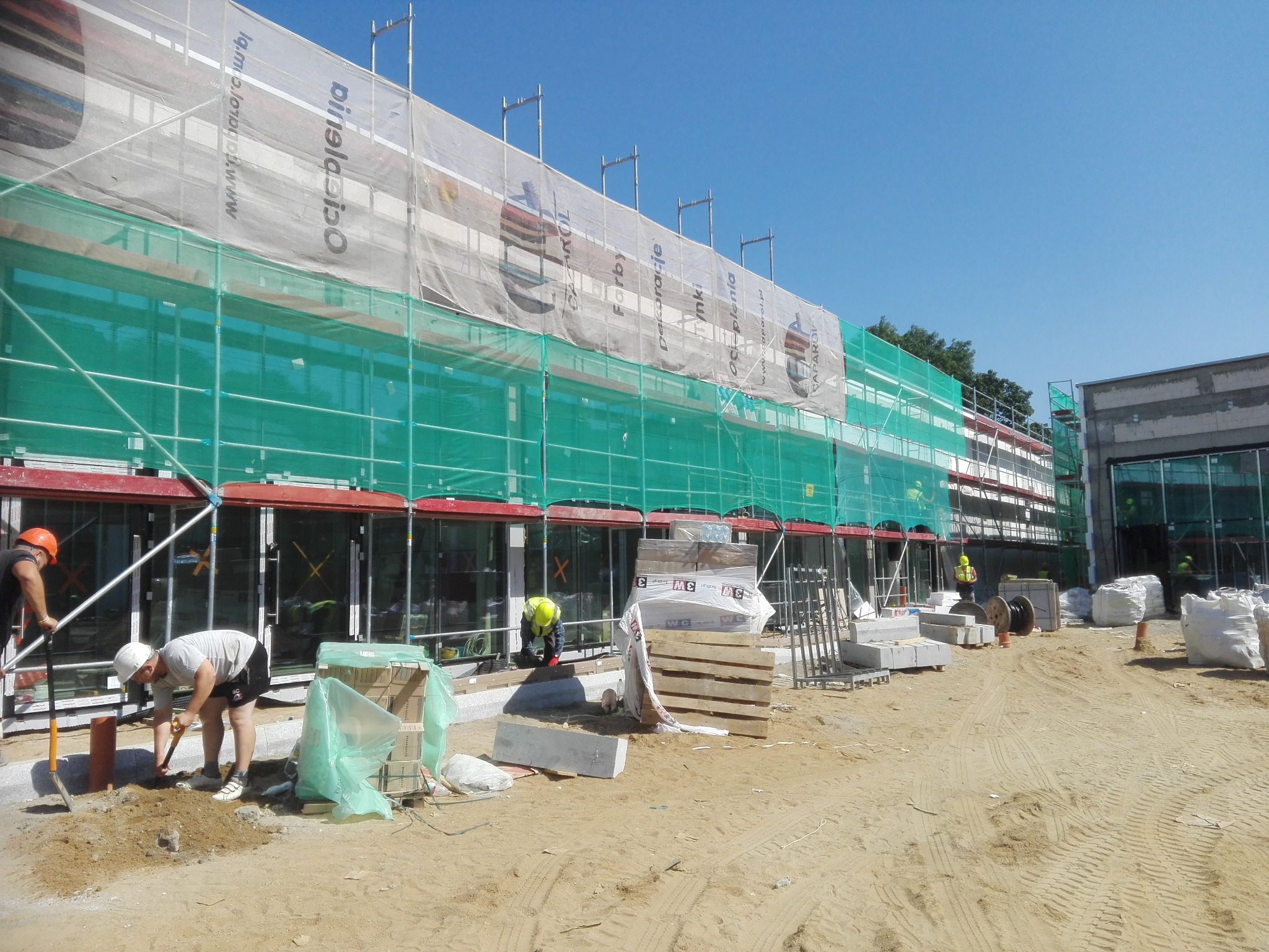 Prace budowlane przy obiekcie z rusztowaniem i siatką ochronną. Widoczni pracownicy w kaskach, materiały budowlane i narzędzia na placu budowy w słoneczny dzień.