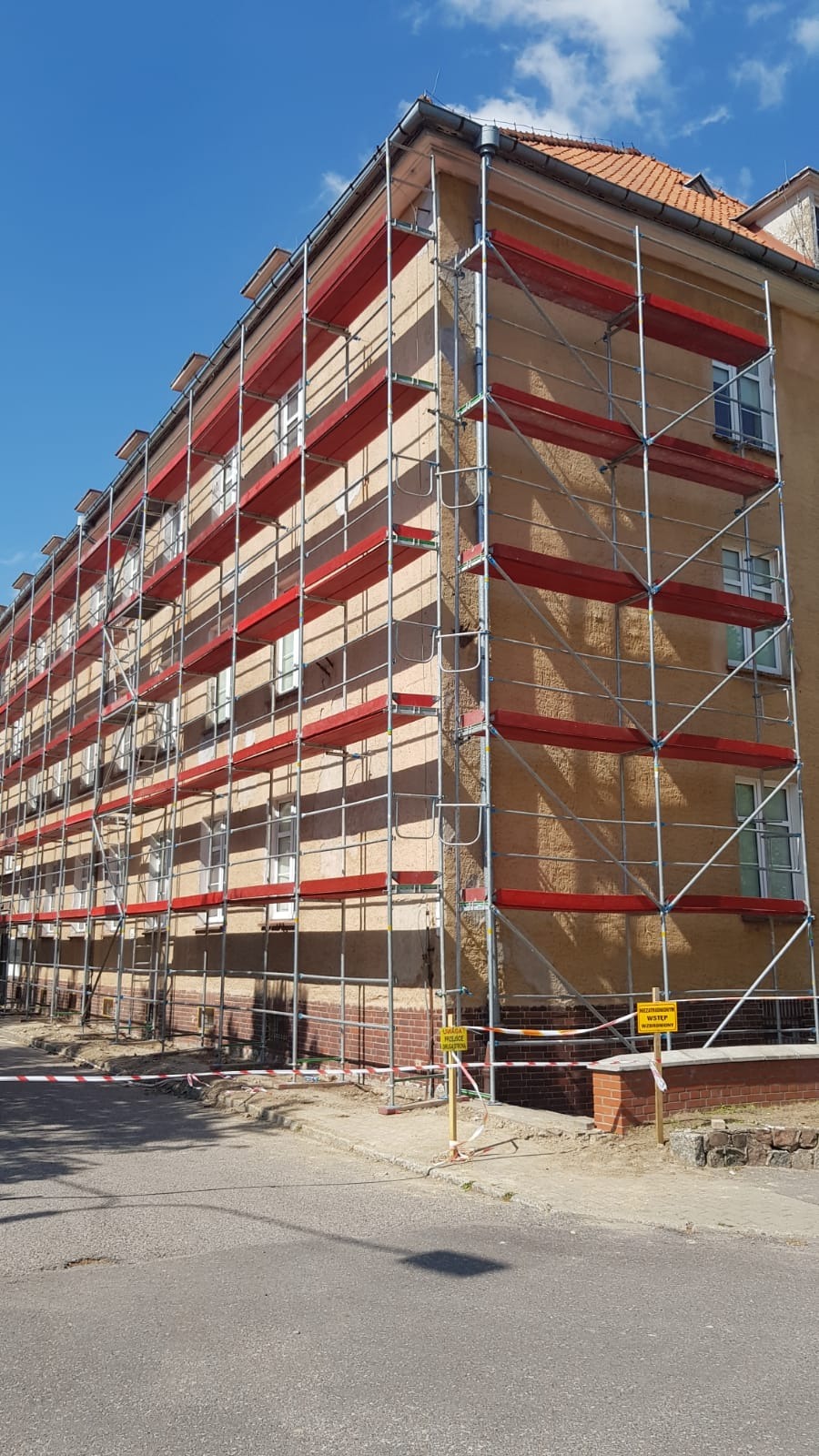 Budynek w rusztowaniach z czerwonymi elementami poziomymi, zabezpieczony taśmą ostrzegawczą i znakami. Prace remontowe lub budowlane w toku, widok z zewnątrz.