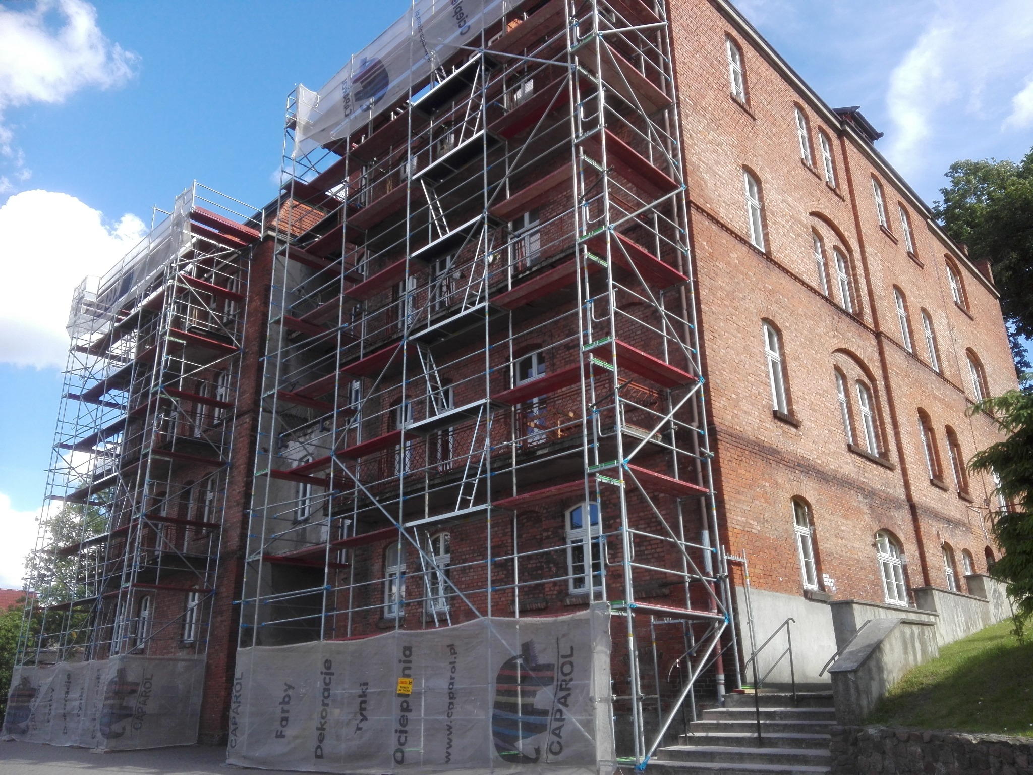 Ceglany budynek w trakcie renowacji, osłonięty rusztowaniami z siatką ochronną z logo Caparol, widok z dołu, błękitne niebo w tle.