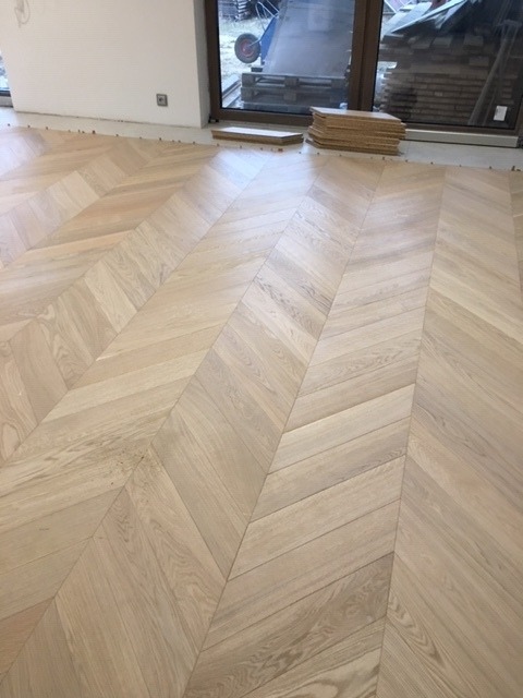 Ułożony parkiet w jodełkę z jasnego drewna, widoczne kliny dystansowe przy ścianie, okno z widokiem na zewnątrz, stos desek parkietowych w tle.