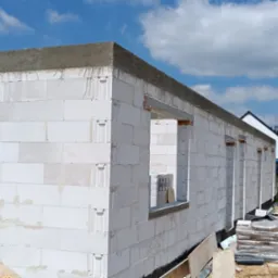 Wznoszenie ścian z białych bloczków betonu komórkowego. Widoczne nadproża okienne, zaprawa na górnej krawędzi muru. W tle błękitne niebo z chmurami i zieleń drzew.