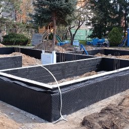 VAH BUD:BUDUIEMO NA VIKY - Fundamenty domu w trakcie izolacji. Widoczne bloczki fundamentowe, zbrojenie i czarna folia izolacyjna. Jeden pracownik montuje izolację. Na drugim planie drzewa.