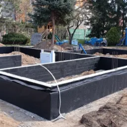 Fundamenty domu w trakcie izolacji. Widoczne bloczki fundamentowe, zbrojenie i czarna folia izolacyjna. Jeden pracownik montuje izolację. Na drugim planie drzewa.