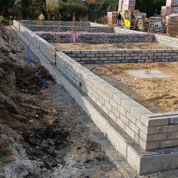 VAH BUD:BUDUIEMO NA VIKY - Budowa fundamentów z cegły klinkierowej w Oławie. Widoczne pręty zbrojeniowe i częściowo ułożona izolacja termiczna. W tle domy jednorodzinne i sprzęt budowlany.