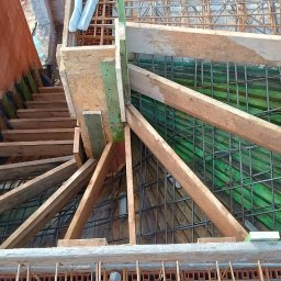 VAH BUD:BUDUIEMO NA VIKY - Z góry widok na szalunek zbrojenia fundamentów z drewnianymi podporami i częściowo wymurowaną ścianą. Widoczne siatki zbrojeniowe i deskowanie.