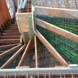 Z góry widok na szalunek zbrojenia fundamentów z drewnianymi podporami i częściowo wymurowaną ścianą. Widoczne siatki zbrojeniowe i deskowanie.