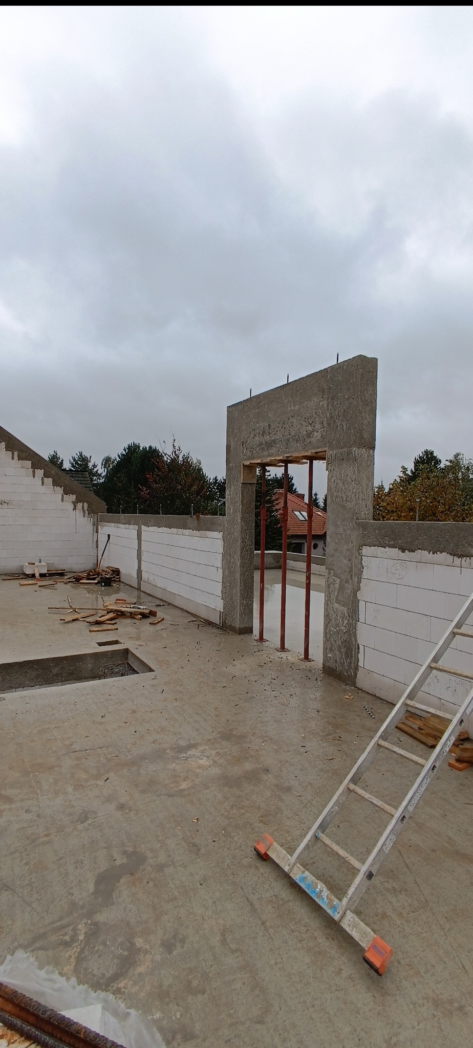 Wnętrze w trakcie budowy: ściany z białych bloczków, betonowy strop z otworem, podpory i drabina. Widok na zewnątrz, pochmurne niebo.