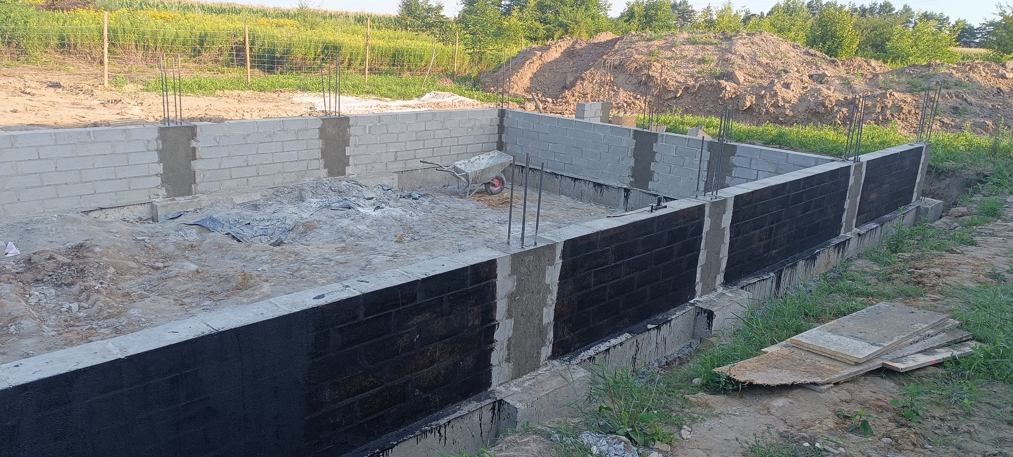 Częściowo ukończone fundamenty z izolacją przeciwwilgociową, widoczne zbrojenia i bloczki betonowe. Na ziemi leżą płyty, w tle zielona łąka.