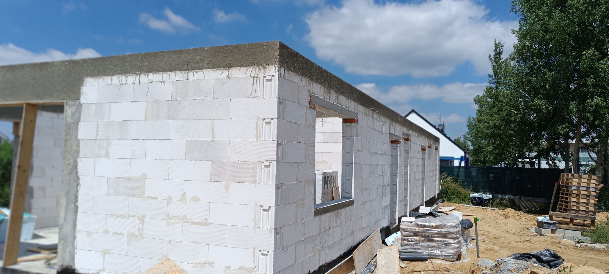 Wznoszenie ścian z białych bloczków betonu komórkowego. Widoczne nadproża okienne, zaprawa na górnej krawędzi muru. W tle błękitne niebo z chmurami i zieleń drzew.