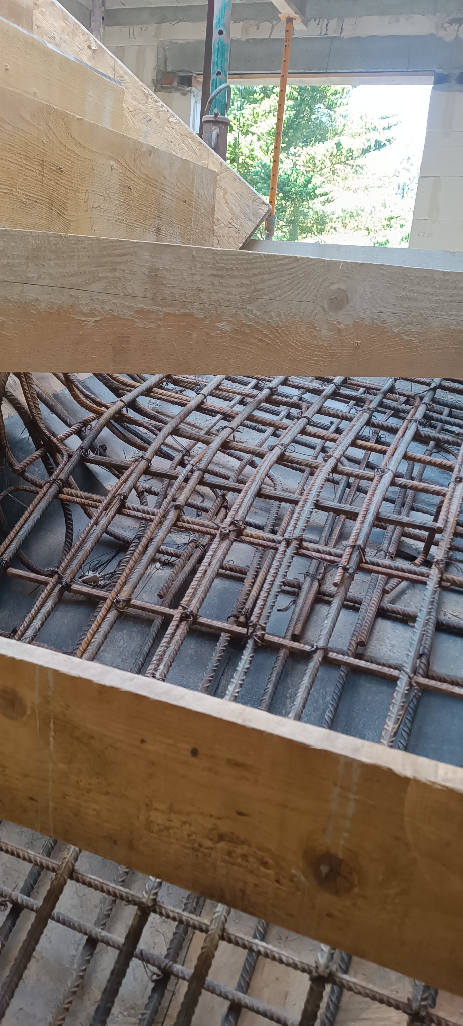 Zbrojenie stropu z widocznymi deskami szalunkowymi i fragmentem ściany z betonu komórkowego w tle. Widok z góry na siatkę z prętów.