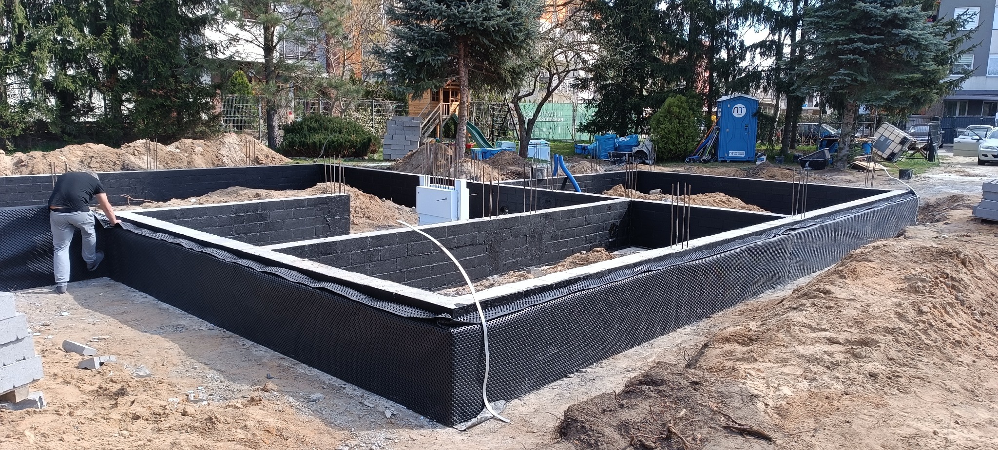 Fundamenty domu w trakcie izolacji. Widoczne bloczki fundamentowe, zbrojenie i czarna folia izolacyjna. Jeden pracownik montuje izolację. Na drugim planie drzewa.