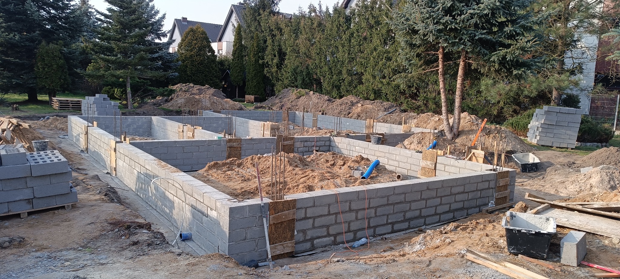 Fundamenty domu z bloczków betonowych w trakcie budowy, widoczne zbrojenia i szalunki, teren budowy z drzewami w tle.