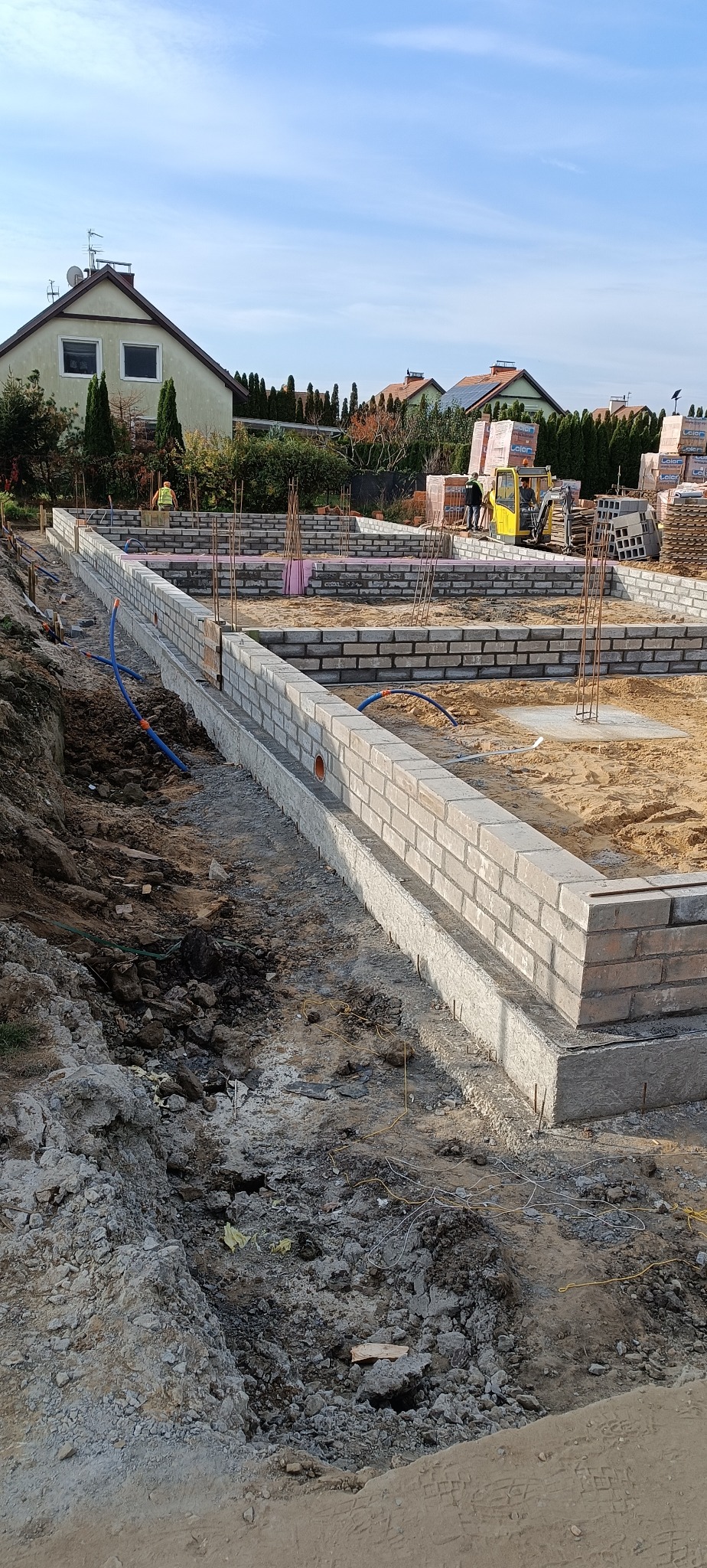 Budowa fundamentów z cegły klinkierowej w Oławie. Widoczne pręty zbrojeniowe i częściowo ułożona izolacja termiczna. W tle domy jednorodzinne i sprzęt budowlany.