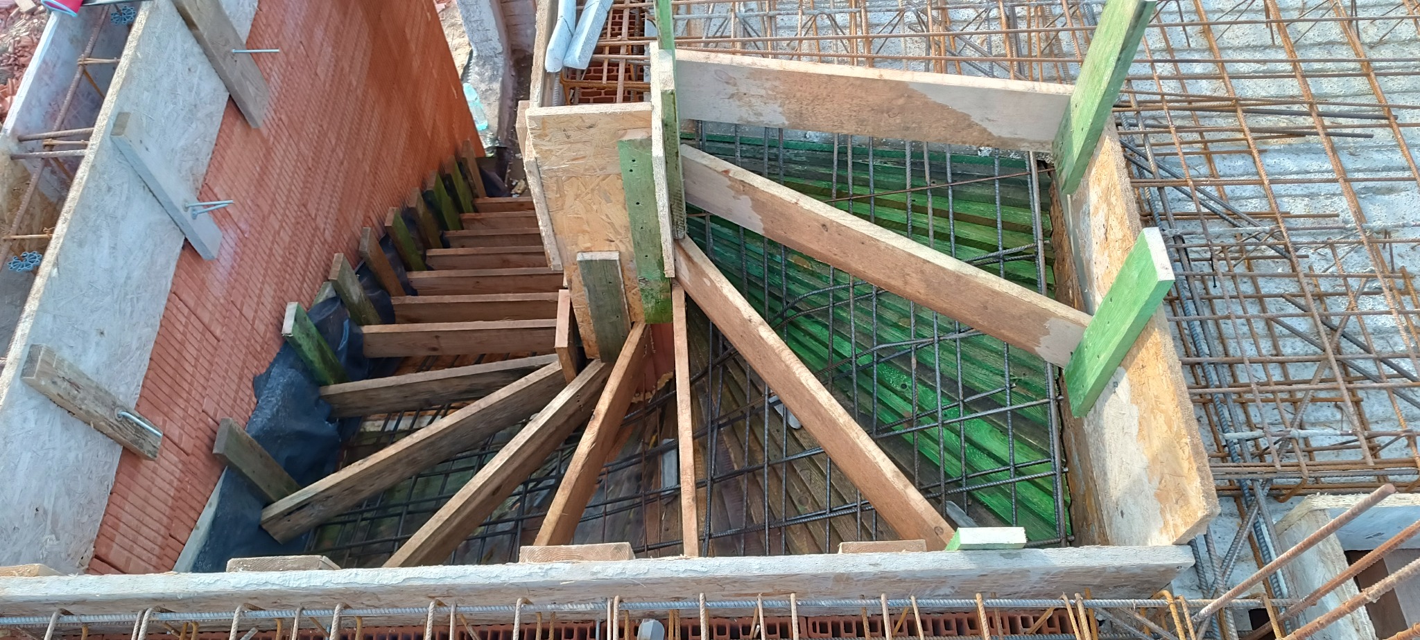 Z góry widok na szalunek zbrojenia fundamentów z drewnianymi podporami i częściowo wymurowaną ścianą. Widoczne siatki zbrojeniowe i deskowanie.