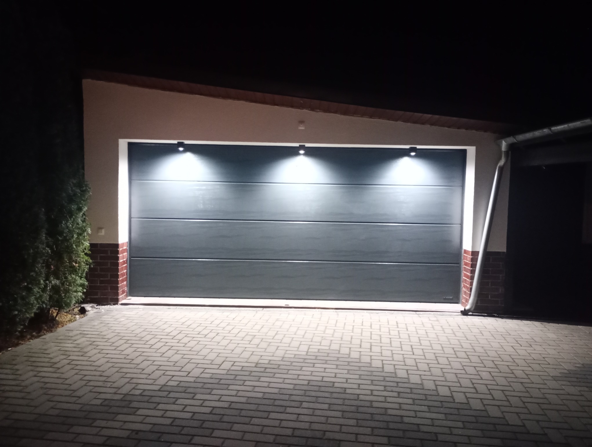 Zamknięta, segmentowa brama garażowa w kolorze grafitowym, oświetlona trzema punktowymi lampami. Podjazd wyłożony kostką brukową. Nocne oświetlenie zewnętrzne.