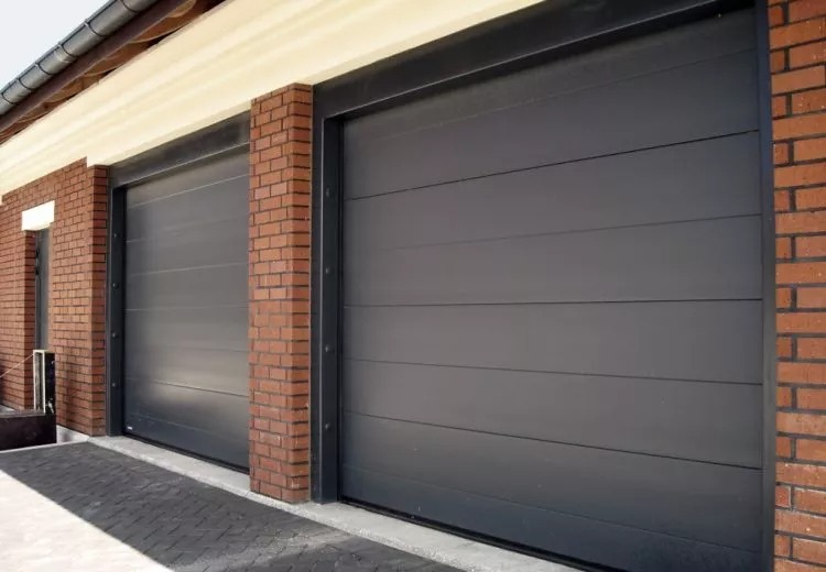 Dwie szare bramy garażowe segmentowe z ceglanymi filarami po bokach, z poziomymi przetłoczeniami, zamontowane w budynku z jasną elewacją i brukowanym podjazdem.