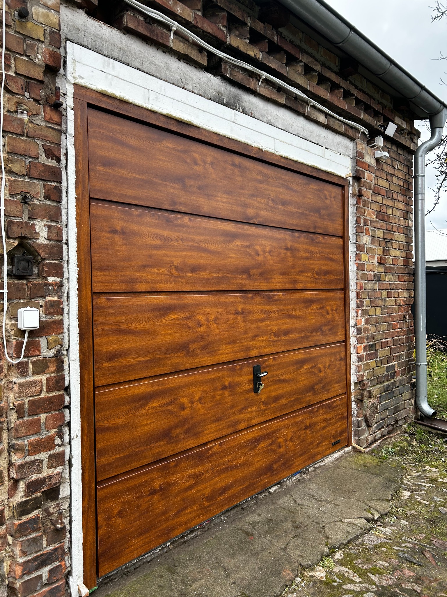 Zamknięta, segmentowa brama garażowa w kolorze drewna, zamontowana w murze z cegły. Widoczne uszczelnienia i elementy montażowe. Ujęcie z dołu, lekko z boku.
