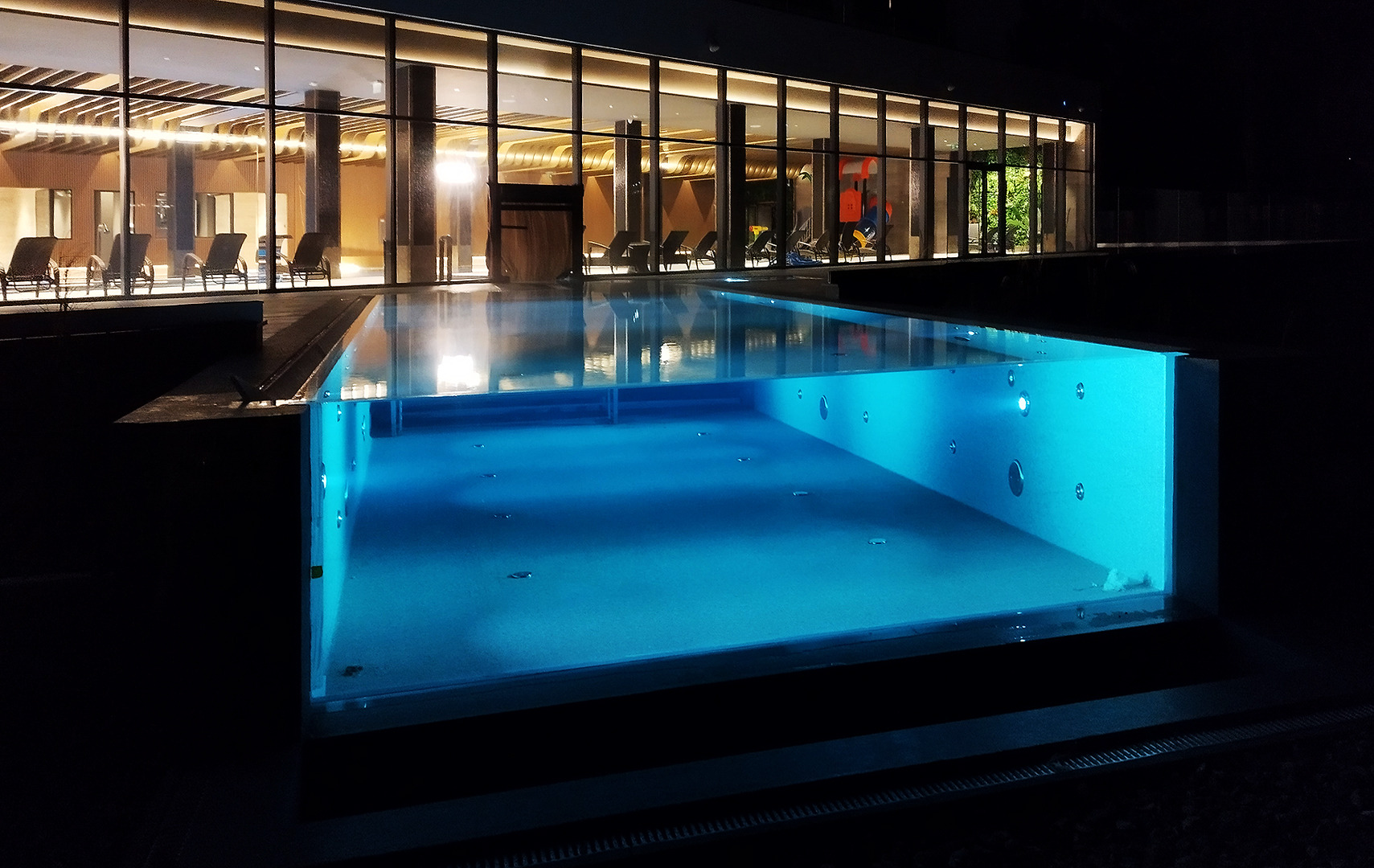 Nocne ujęcie podświetlonego, nowoczesnego basenu z jacuzzi, z widokiem na przeszklony budynek z leżakami wewnątrz. Minimalistyczny design, ciemne otoczenie.