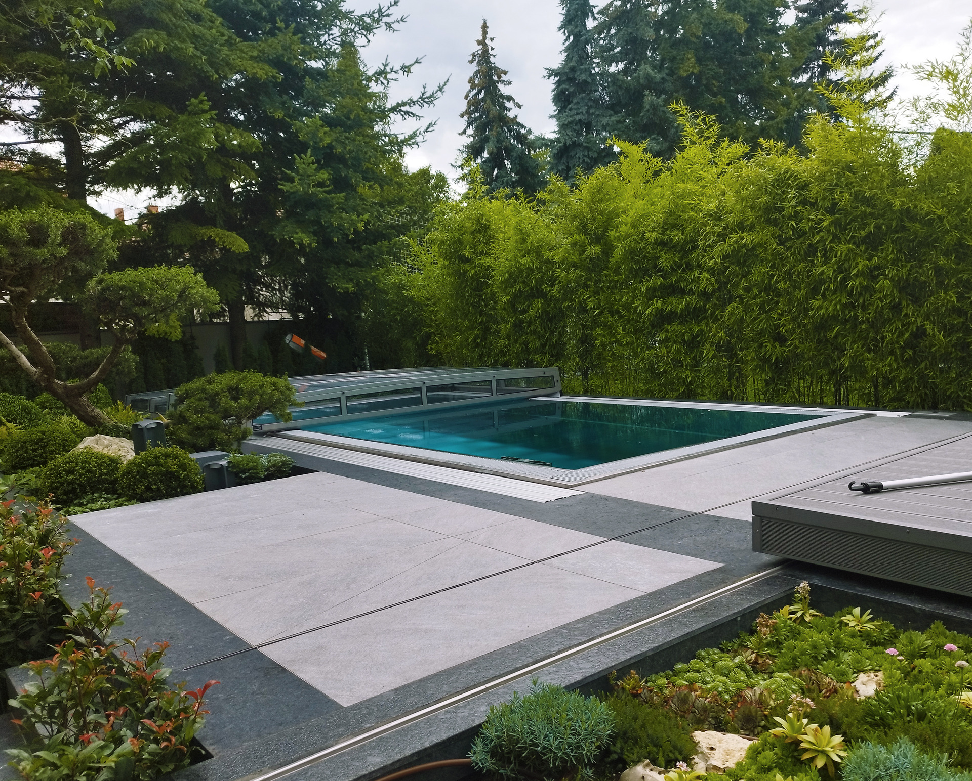Nowoczesny basen otoczony szarą, kamienną nawierzchnią i zielenią. Widoczne bambusy i drzewa w tle. Częściowo zasłonięta pokrywa basenu.