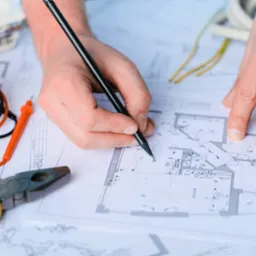 Ręka kreśląca plan instalacji elektrycznej na projekcie architektonicznym; obok miernik, kombinerki i przewody. Plan oświetlenia w jasnym pomieszczeniu.