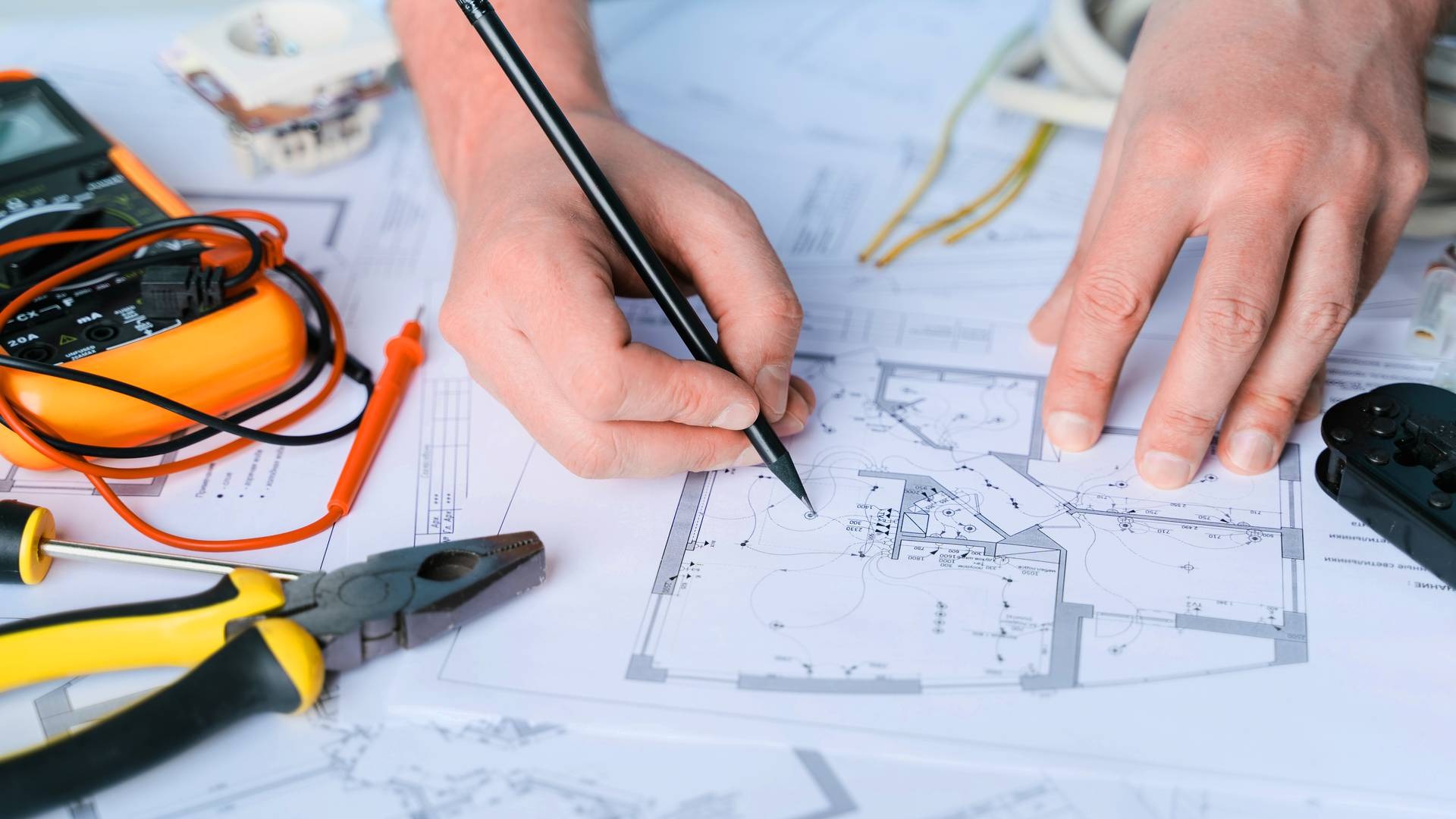 Ręka kreśląca plan instalacji elektrycznej na projekcie architektonicznym; obok miernik, kombinerki i przewody. Plan oświetlenia w jasnym pomieszczeniu.