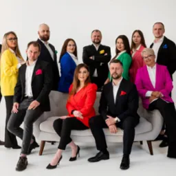 Profesjonalny zespół nieruchomości w kolorowych marynarkach pozuje na tle białej ściany, siedząc na szarej sofie. Elegancki ubiór i uśmiechy budują wizerunek zaufania.