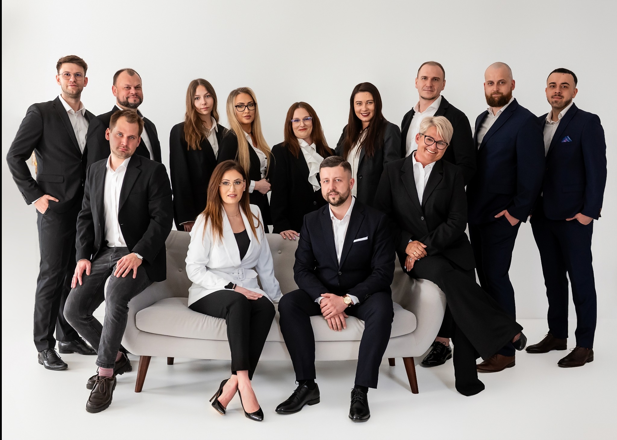 Zespół biura nieruchomości pozuje w garniturach na jasnym tle. Kobiety w białych koszulach i marynarkach siedzą na sofie, mężczyźni stoją za nimi.