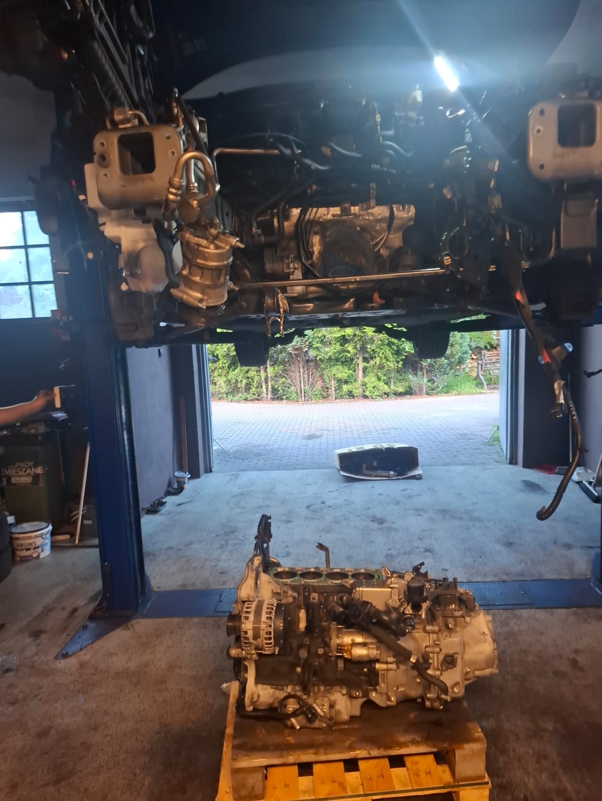 Silnik samochodowy wyjęty z pojazdu, na palecie w warsztacie. Auto na podnośniku, widoczne elementy podwozia. Otwarta brama garażowa w tle.