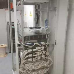 Szafa serwerowa z zainstalowanymi urządzeniami sieciowymi i uporządkowanymi kablami, widok z przodu, w tle biała ściana.