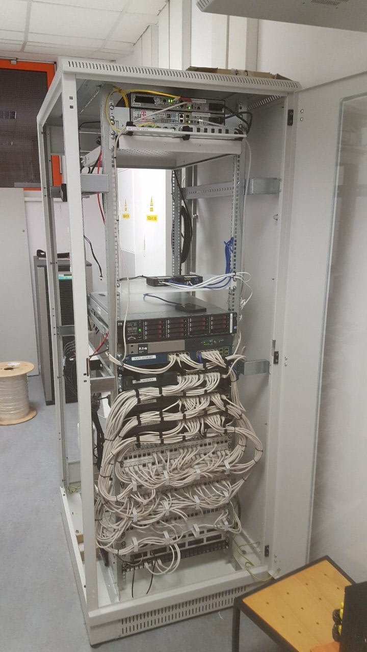 Szafa serwerowa z zainstalowanymi urządzeniami sieciowymi i uporządkowanymi kablami, widok z przodu, w tle biała ściana.