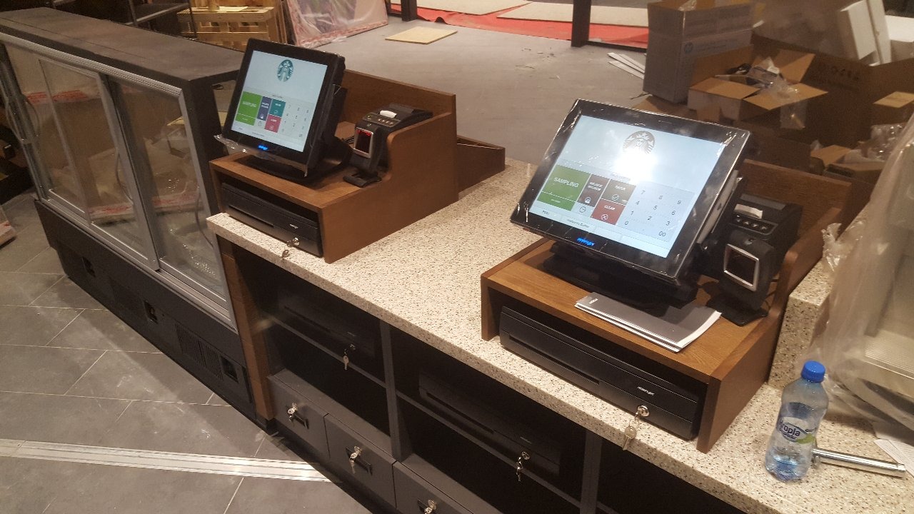 Dwa stanowiska kasowe z ekranami dotykowymi w trakcie instalacji, widoczne kasy fiskalne, czytniki kart i butelka wody. Tło stanowią materiały budowlane.