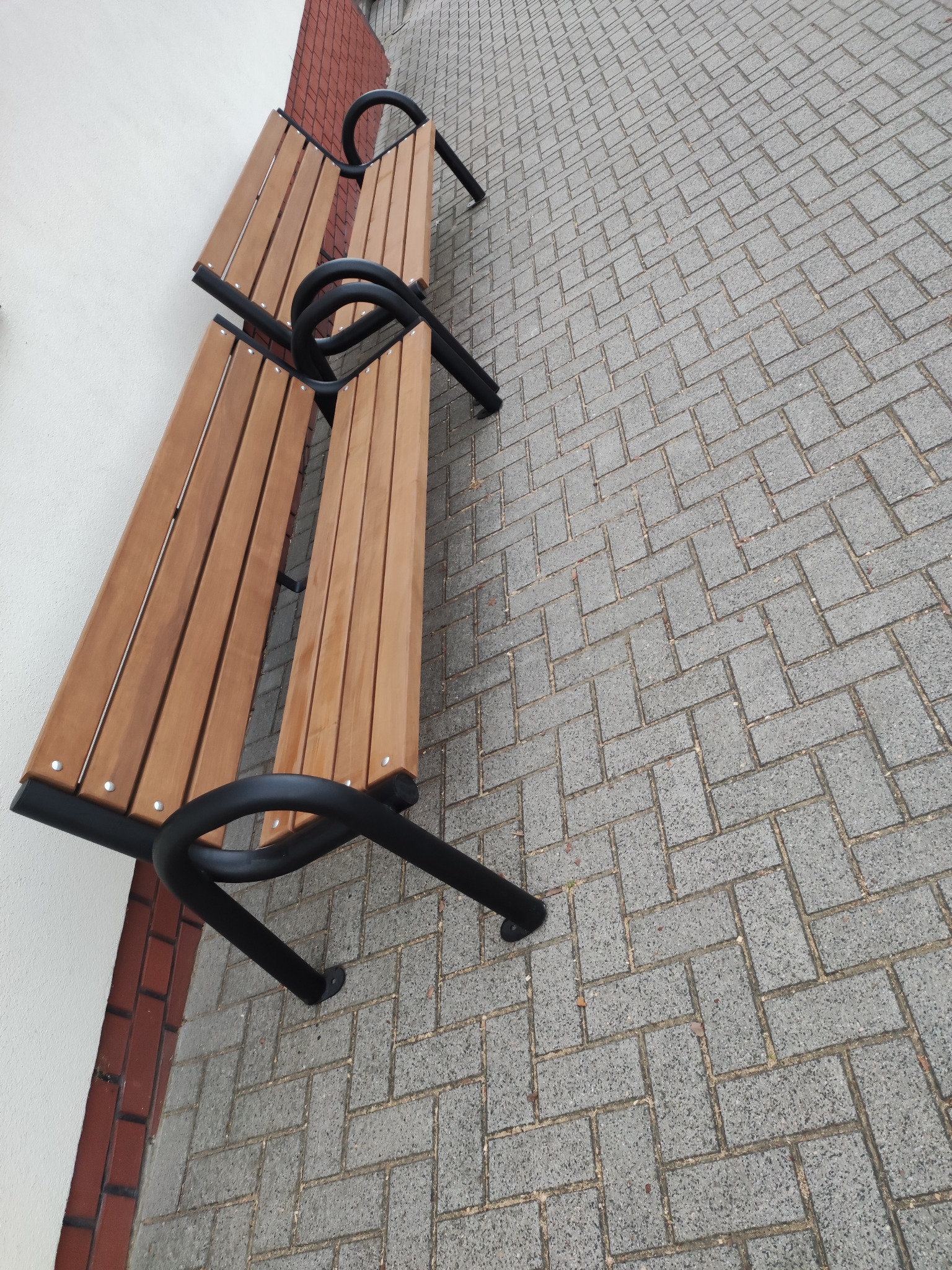 Dwie drewniane ławki z czarnymi metalowymi nogami, ustawione na szarym chodniku z kostki brukowej, widoczne z góry pod kątem.