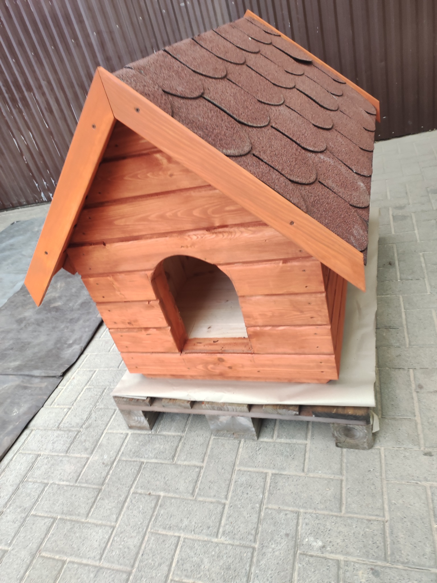 Drewniana buda dla psa z brązowym dachem z gontu bitumicznego, ustawiona na palecie. Widoczna tekstura drewna i precyzyjne wykonanie.