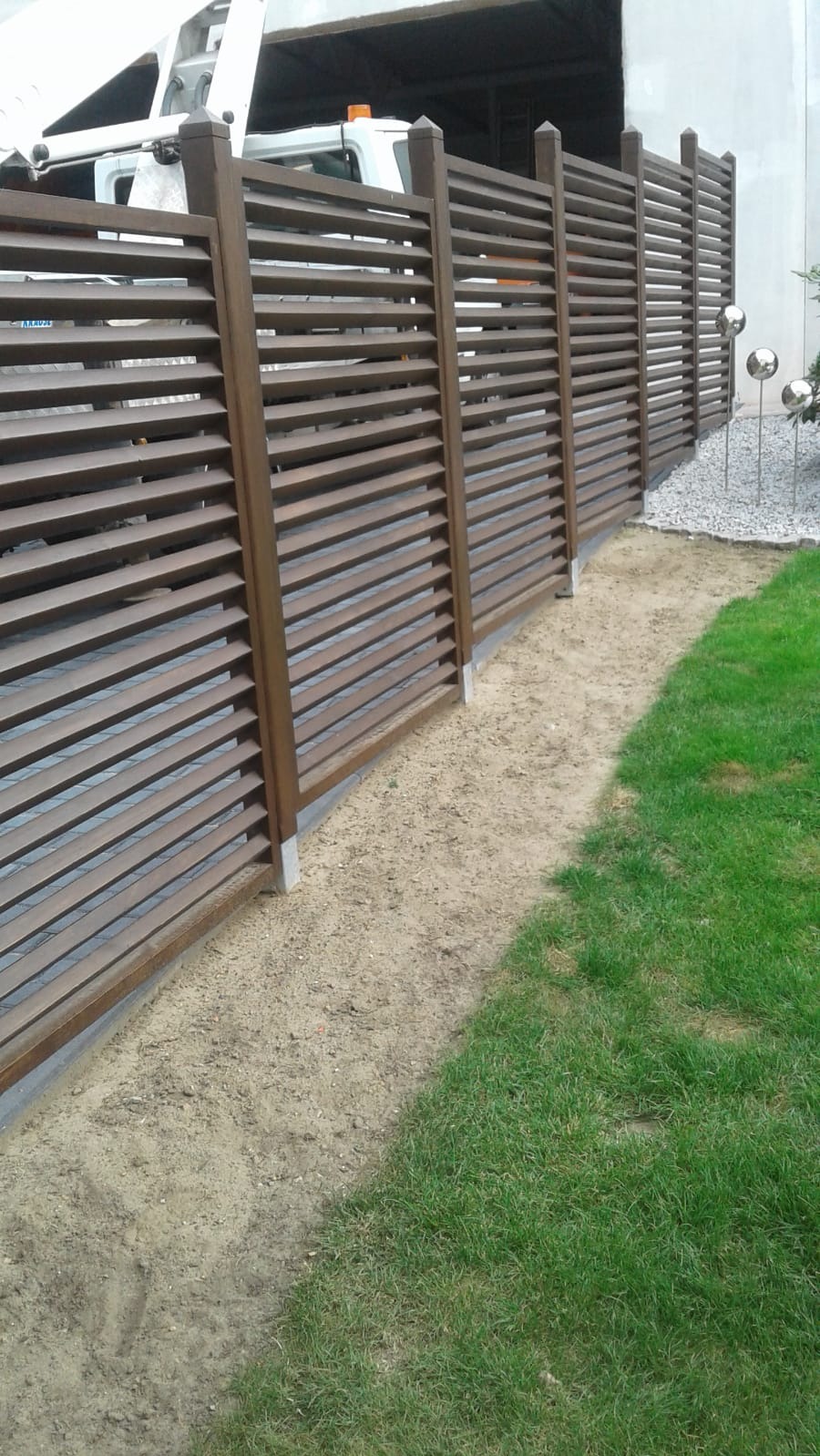 Nowoczesne, poziome ogrodzenie lamelowe w kolorze brązowym, oddzielające trawnik od żwirowego podłoża. W tle zaparkowany samochód dostawczy.
