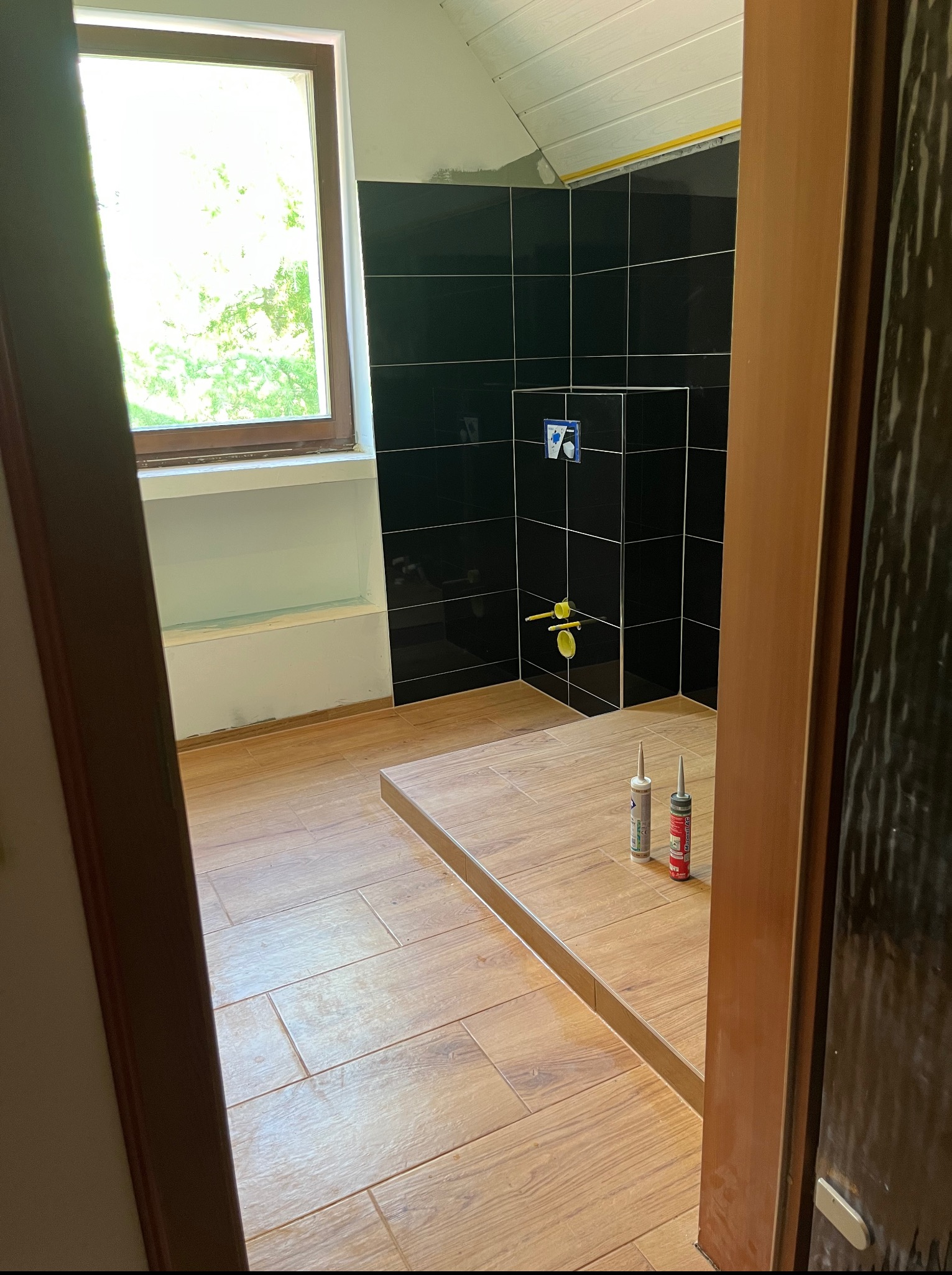 Częściowo wykończona łazienka z czarnymi płytkami na ścianach, drewnopodobnymi płytkami na podłodze i oknem z widokiem na zieleń. Widoczne narzędzia do montażu.