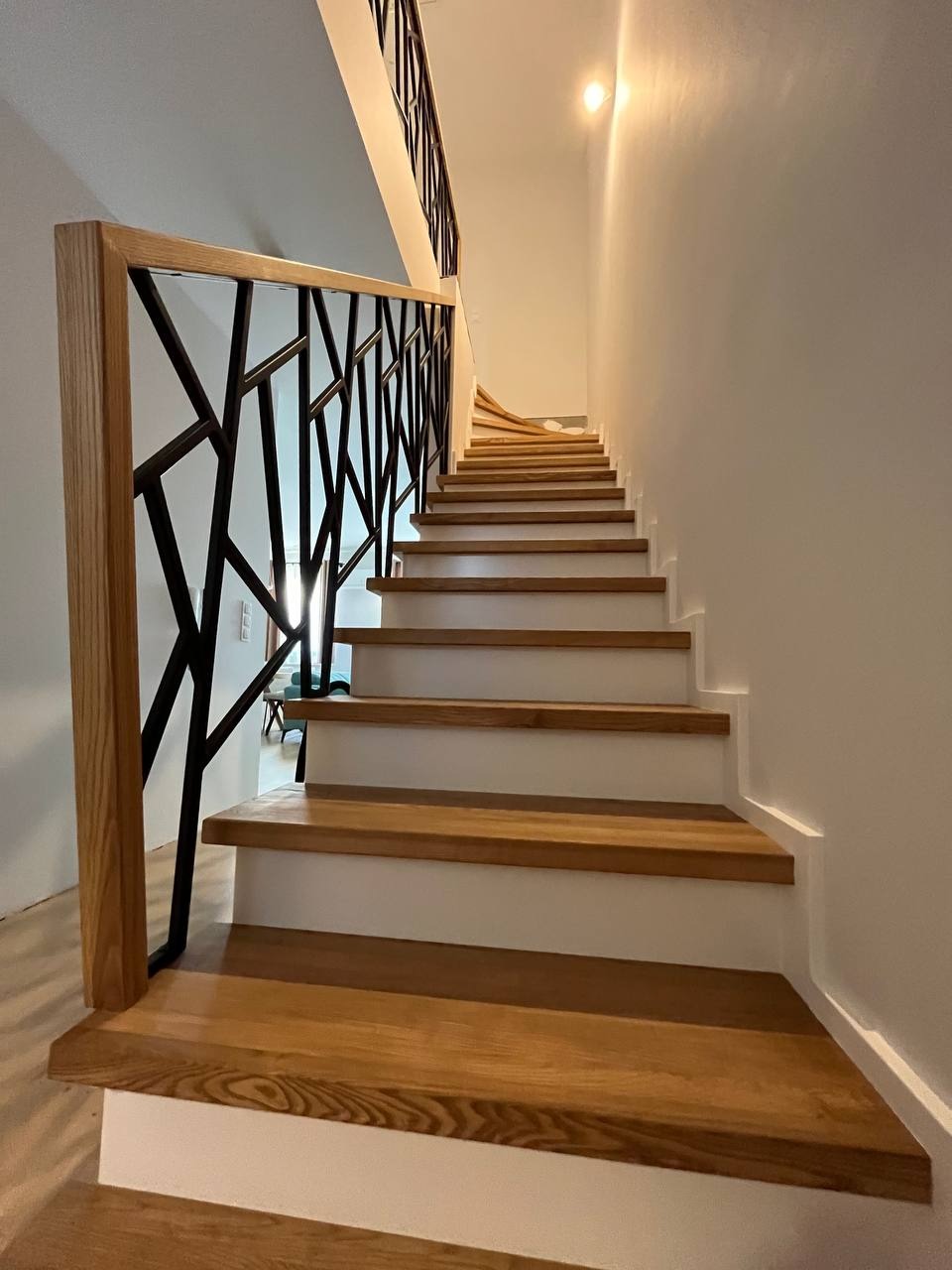 Nowoczesne schody z drewnianymi stopniami i balustradą z czarnych, geometrycznych elementów na jasnym tle. Minimalistyczny design i ciepłe oświetlenie.