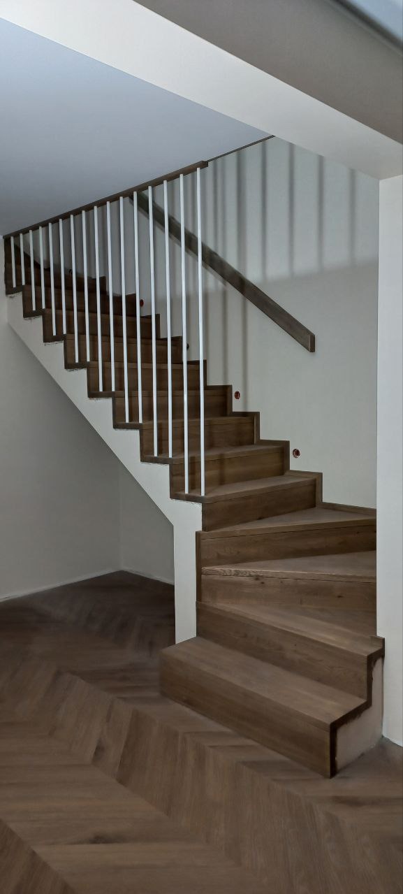 Drewniane schody z białą balustradą na tle jasnej ściany. Widoczne oświetlenie schodów i parkiet w jodełkę. Schody wykonane z drewna o ciepłym odcieniu.