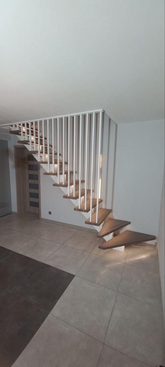 Nowoczesne schody z drewnianymi stopniami i białą, pionową balustradą. Stopnie podświetlone od spodu, jasne płytki podłogowe. Minimalistyczny design.
