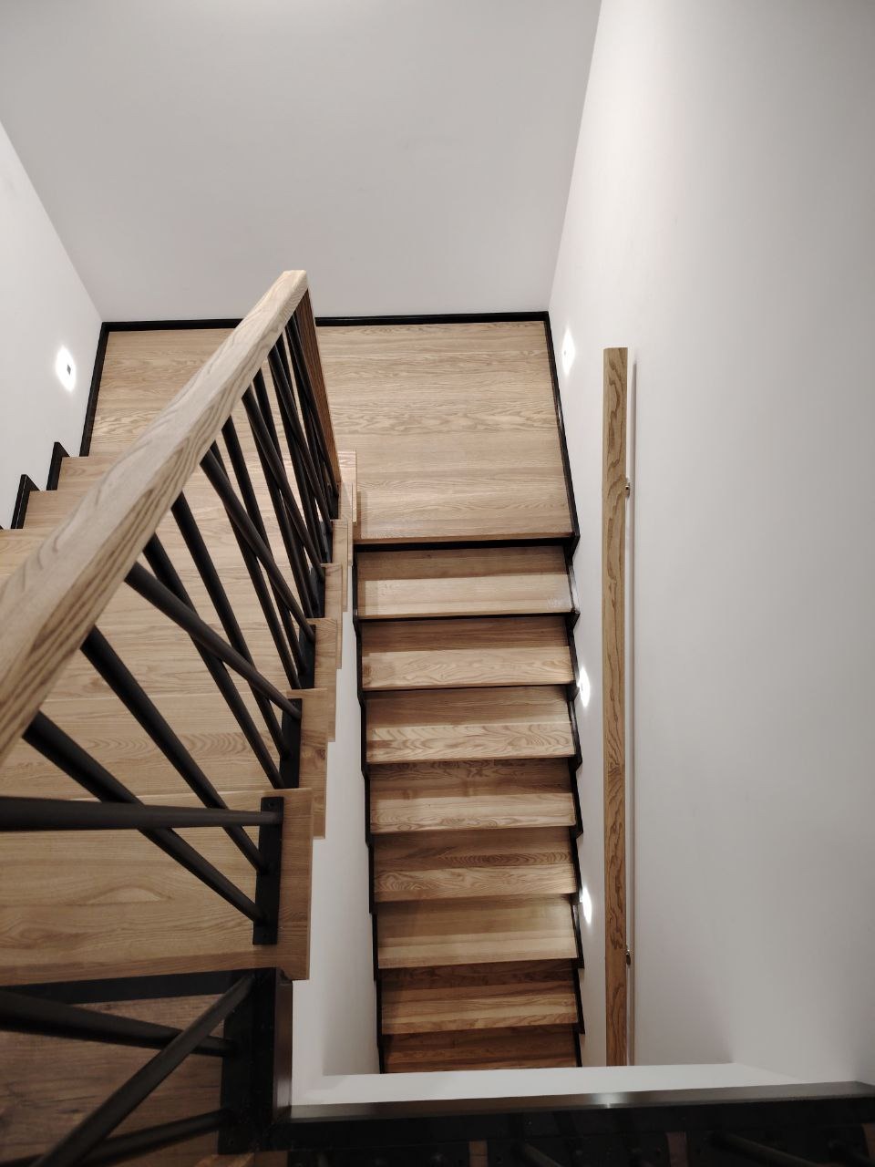 Nowoczesne schody z jasnego drewna z czarnymi, metalowymi elementami balustrady, widziane z góry. Minimalistyczny design, oświetlenie punktowe w ścianie.