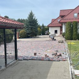 Usługi brukarskie i Ogrodzenia . - Układanie wielobarwnej kostki brukowej na podjeździe domu jednorodzinnego. Widoczny fragment ułożonej nawierzchni i pracujący mężczyzna. W tle dom z czerwoną dachówką.
